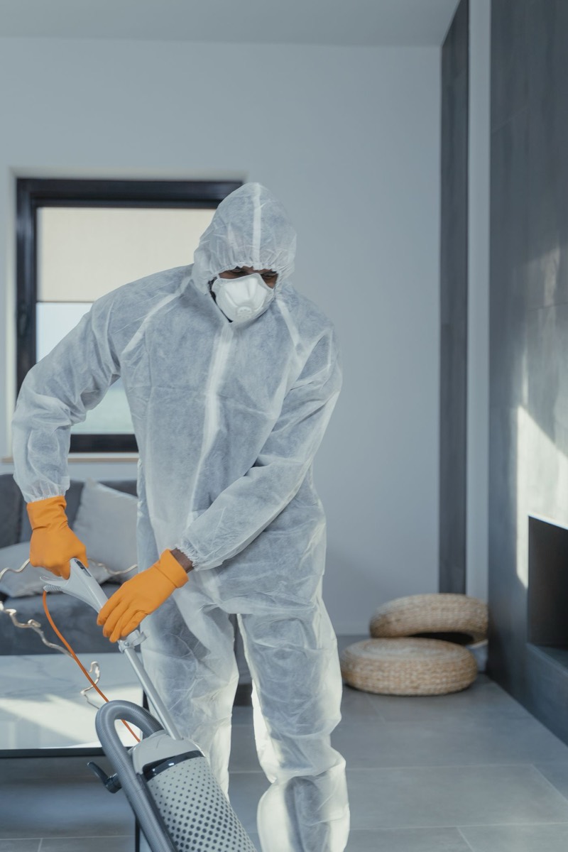 Worker in a white protective suit, mask and gloves using an industrial vacuum indoors